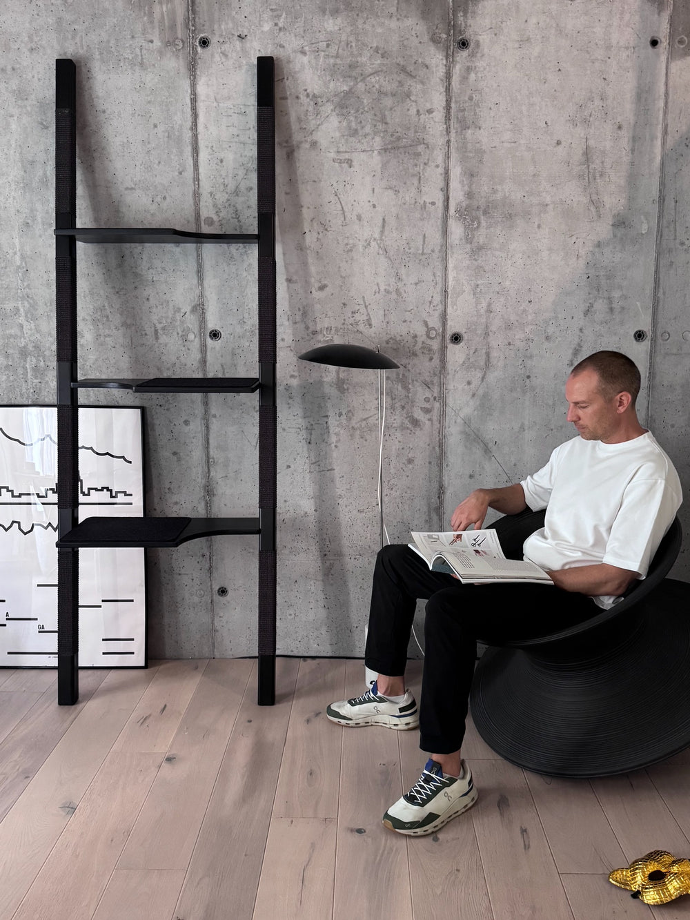 Design-Kratzbaum H-Tree H3 aus lackiertem Echt-Holz in Schwarz mit schwarzem Sisal vor Betonwand – modernes Wohnzimmer mit minimalistischer Einrichtung, Mann liest entspannt im Stuhl daneben