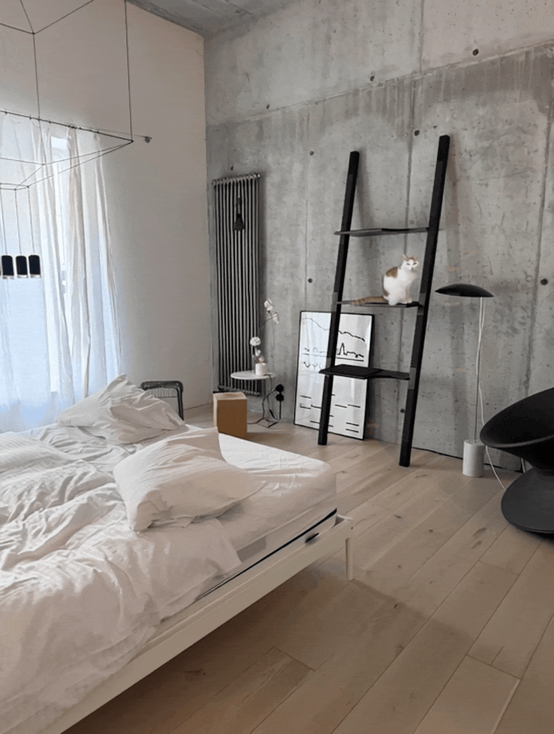 Softly lit bedroom with an unmade white duvet and a cat perched on ablack H-Tree cat tree nearby.