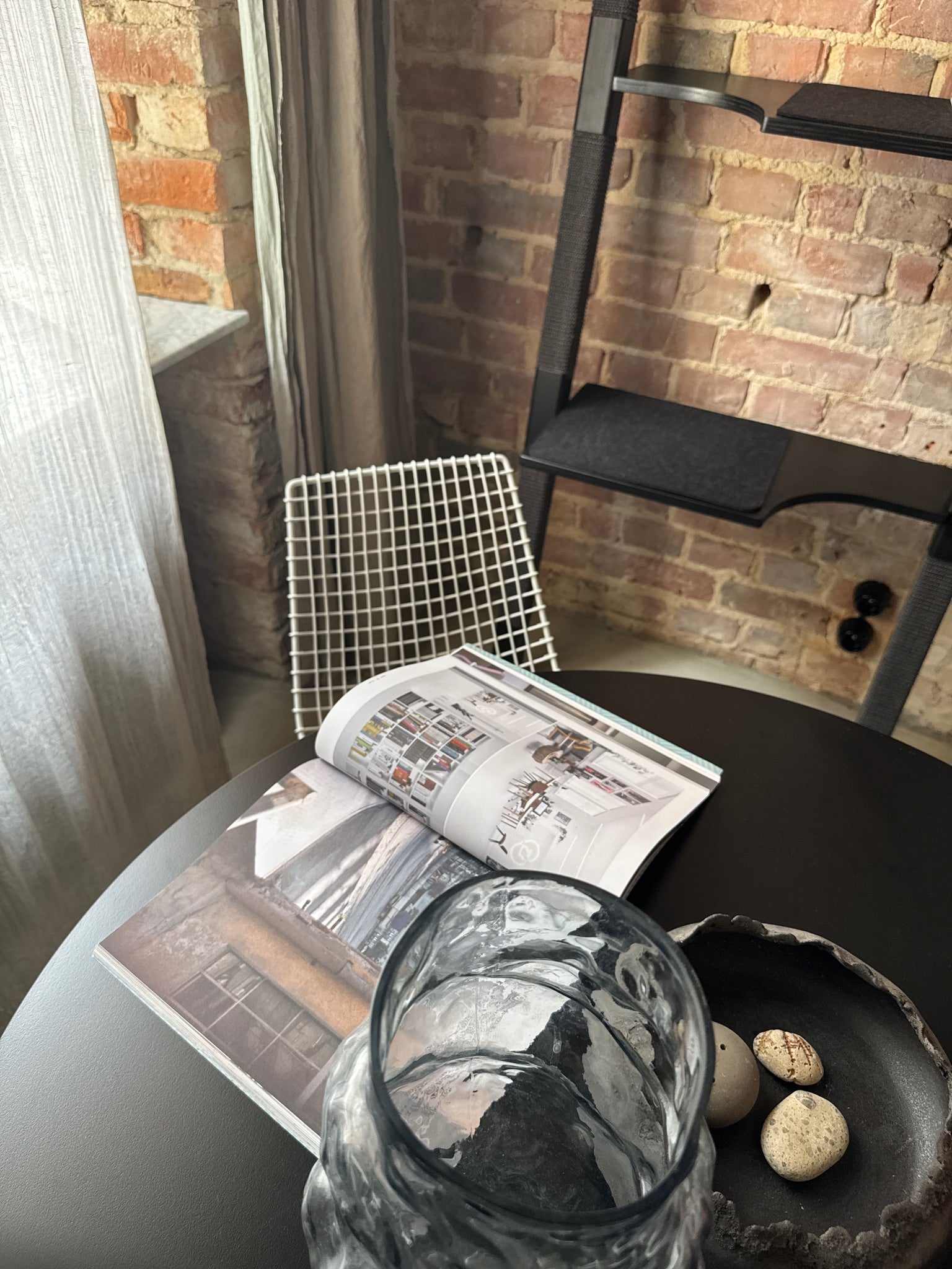 Table with an open interior design magazine, glass vase, and decorative bowl in the foreground, with a black H-Tree cat ladder standing against a brick wall in the background.